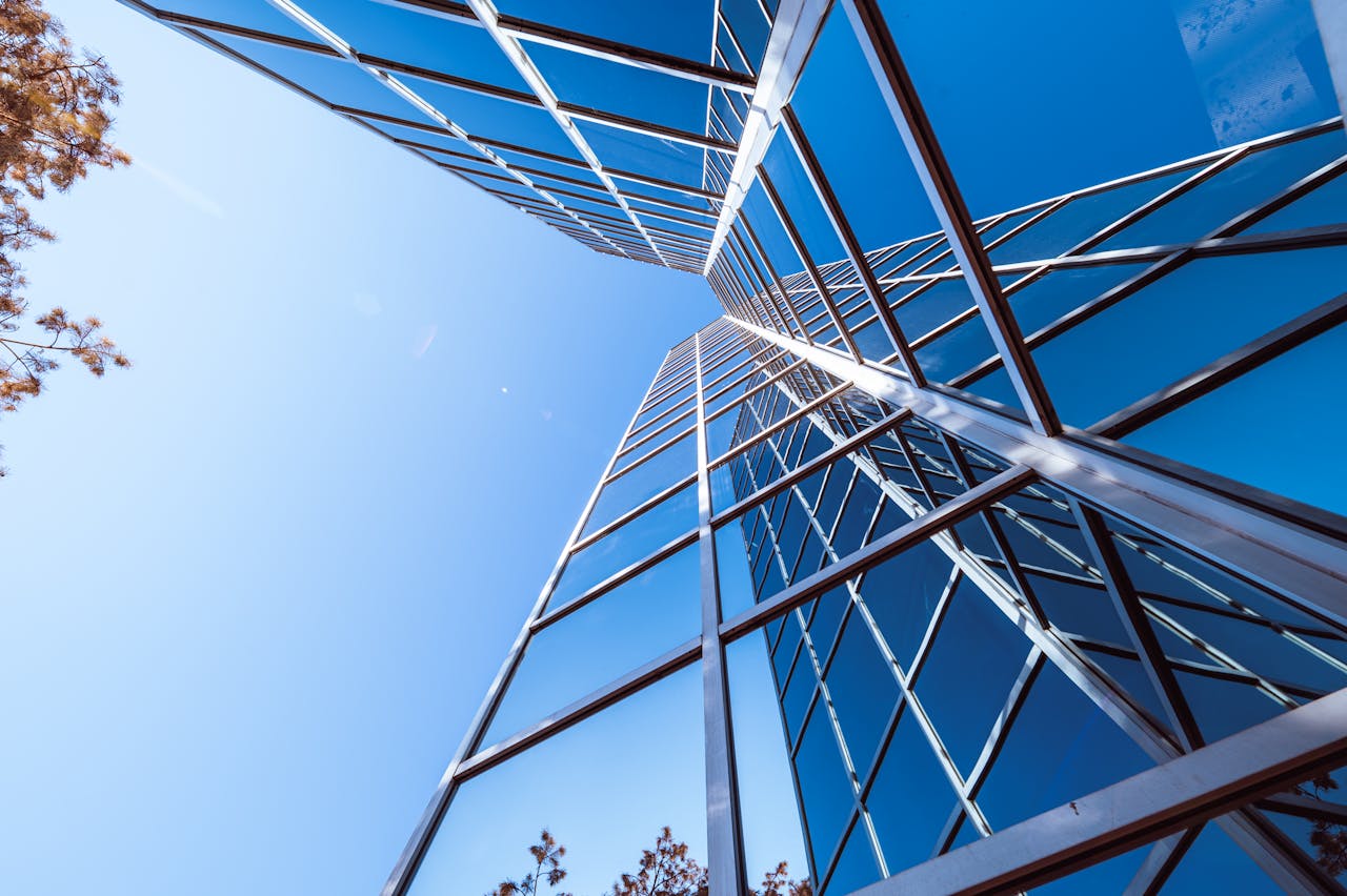 Low angle of glass mirrored facade of contemporary multistory building of unusual shape reflecting cloudless blue sky and tree on sunny day